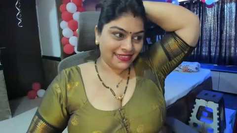 Media: Video of a smiling South Asian woman with medium skin tone, wearing a green blouse, gold necklace, and red bindi. She poses confidently indoors with a festive backdrop, featuring red and white balloons and a decorative curtain.