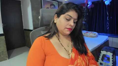 Media: Video of a South Asian woman with long black hair, wearing a bright orange sari, sitting at a desk in a modern office with dark curtains and colorful balloons.