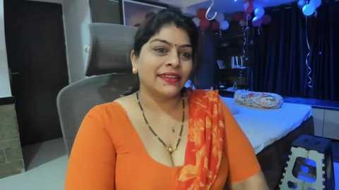 Media: Video of a South Asian woman with dark hair, medium skin tone, wearing an orange sari and gold necklace, seated in a modern room with a beige chair, white table, and blue balloons.