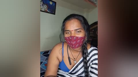 Media: Video of a woman with medium skin tone and long black hair, wearing a blue bra, red mask, and a black-and-white striped top. She sits in a room with a blue geometric-patterned cushion, a framed image, and a wooden cabinet in the background.