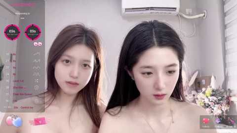 Media: Video of two East Asian women with fair skin, long black hair, and small breasts, posing topless in a modern, minimalist bedroom with white walls and a TV screen showing a live stream.