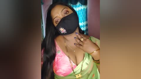 Media: Video of a South Asian woman with medium-dark skin, wearing a black mask, pink and green saree, and gold accessories, standing in a dimly lit room.