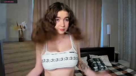 Media: A video of a young woman with curly brown hair and fair skin wearing a white sports bra with numerical patterns, standing in a modern bedroom with beige curtains and a wooden dresser.