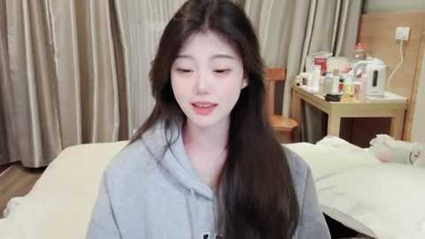 Media: Video of an Asian woman with long black hair, wearing a gray robe, sitting on a bed in a hotel room with beige curtains, wooden furniture, and toiletries on a nightstand.