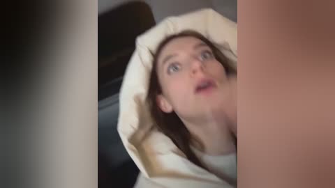 Media: Video of a pale-skinned, fair-haired woman with wide eyes and open mouth, wearing a beige blanket, with blurred, dark-haired man in the background.