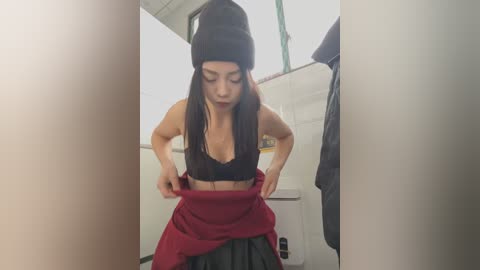 Media: Video of a young woman with long dark hair, wearing a black beanie, black bra, and red skirt, adjusting her clothing in a dimly lit bathroom.