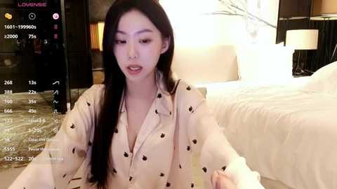 Media: Video of a young East Asian woman with long black hair, wearing a white blouse with black buttons, sitting on a bed in a modern bedroom.