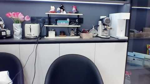 Media: Video of a modern kitchen counter with white cabinets, black countertops, and a black chair in the foreground. The counter holds an electric kettle, coffee maker, decorative flowers, and a white tray with a blue box.