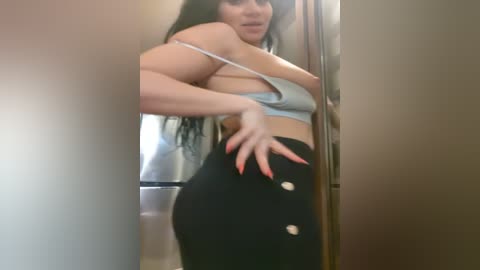 Media: A video of a curvy woman with long black hair, wearing a light blue crop top and black high-waisted pants with silver buttons, posing provocatively in a dimly lit room.