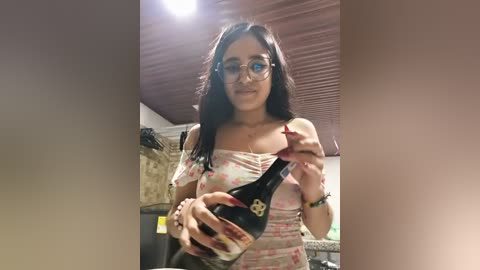 Media: Video of a young woman with long black hair, wearing glasses, a floral off-shoulder top, and holding a black wine bottle. She's in a dimly lit room with a wooden ceiling and a wall of shelves.