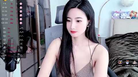 Media: Video of an East Asian woman with long black hair, fair skin, and red lipstick, wearing a sleeveless, patterned top, sitting in a room with a bed, toys, and a recording device.