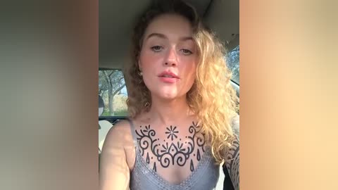 Media: Video of a fair-skinned woman with curly blonde hair, wearing a gray lace bralette, showcasing intricate black floral tattoo on her chest, seated in a car with sunlight streaming in.