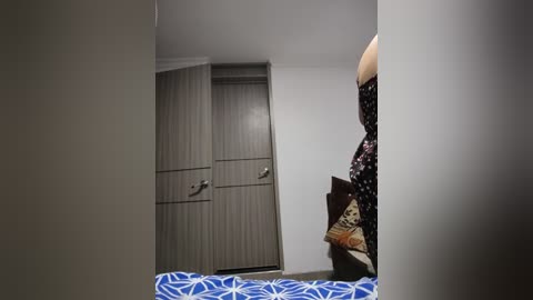 Media: Video of a woman in a black floral dress standing in a minimalist, white-walled room with a blue-patterned bedspread and a closed, grey wooden wardrobe.