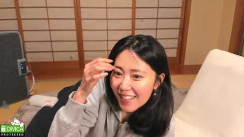 Media: Video of a cheerful East Asian woman with long black hair, wearing a gray hoodie, adjusting her glasses, seated on a beige couch in a traditional Japanese room with wooden screens and tatami mats.