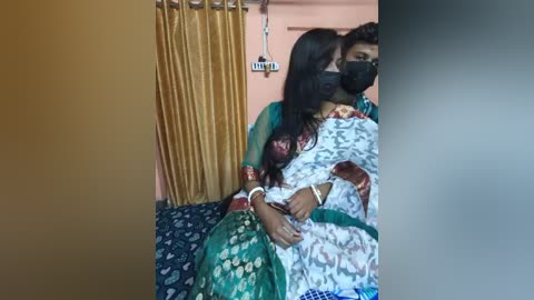 Media: A video shows a woman with long dark hair, wearing a green and white saree, lying on a bed with a blue patterned sheet. She has a mask and a bracelet on her arm.