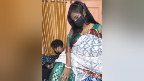 Media: Video of a young woman in a green sari, black mask, and white blouse caring for a child lying on a bed in a room with golden curtains.