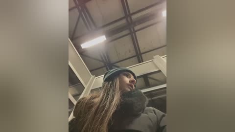 Media: A video captures a young woman with long, dark hair and a green beanie, wearing a dark coat, inside a dimly lit, industrial building with exposed metal beams and a fluorescent light fixture.