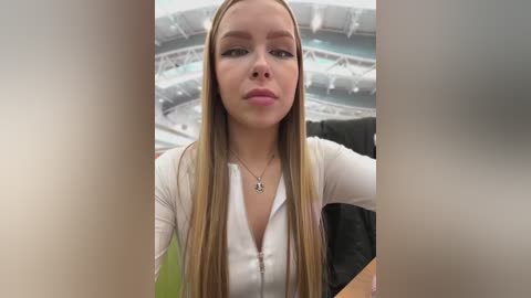 Media: Video of a young Caucasian woman with long blonde hair, wearing a white zip-up top, sitting in a stadium.