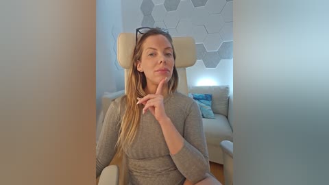 Media: Video of a blonde woman in a grey sweater, sitting on a modern chair, with a thoughtful expression and finger to her lips in a minimalist living room.