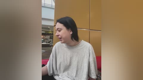 Media: Video of a fair-skinned woman with short black hair, wearing a gray t-shirt, smiling, in a modern, beige-walled room with red furniture and a blurred background of a caf\u00e9.