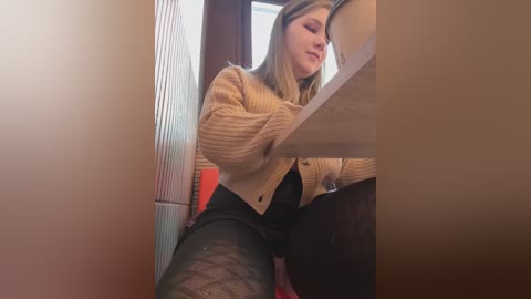 Media: A video of a young Caucasian woman with light skin, blonde hair, and a slim build, wearing a beige knitted cardigan, black lace top, and black tights, sitting at a wooden table in a dimly lit room.