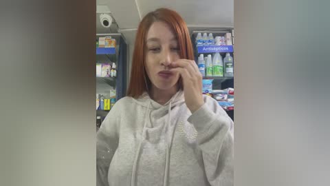 Media: Video of a young woman with long, straight red hair, wearing a light gray hoodie, standing in a brightly lit convenience store, holding a bottle of water.