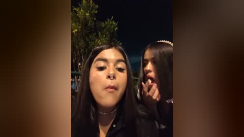 Media: Video of two young women with dark hair, one with a septum piercing, both in dark clothing, standing outdoors at night, with blurred foliage in the background.