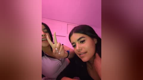 Media: Video of a young Latina woman with medium skin tone, long dark hair, and a nose ring, making a peace sign, standing in a pink-lit room with a mirror reflection of herself.