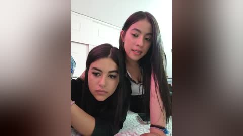 Media: Video of two young Latina women with long dark hair, one in a black top, the other in a white top, sitting closely on a bed with patterned sheets, in a dimly lit room.