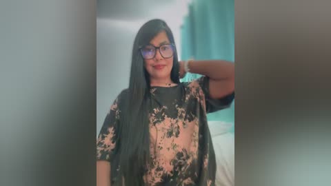 Media: A video of a young woman with long black hair, wearing glasses, a floral blouse, and a black shawl, standing indoors against a teal curtain backdrop.