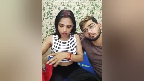 Media: Video of a young South Asian woman with red streaks in her hair, wearing a striped crop top and black pants, seated next to a man with glasses, both holding a red drink in a public bathroom.