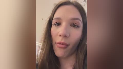 Media: Video of a young Caucasian woman with fair skin, long brown hair, and a neutral expression, standing in a beige room with a wooden door to her right.