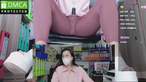 Media: Video of an Asian woman in a pink shirt and pink pants sitting on a desk in a cluttered office, wearing a face mask, with a \"DMCA Protected\" watermark.