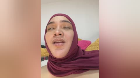 Media: Video of a young woman with medium skin tone, wearing a burgundy hijab and a mustard-yellow sweater, lying on a bed with a pink pillow.