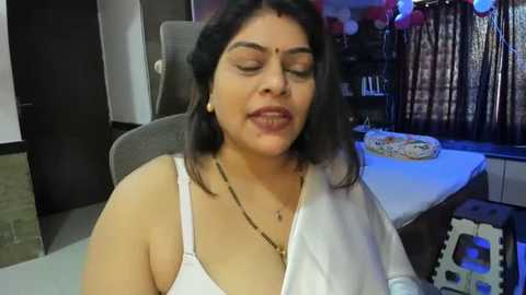 Media: Video of a South Asian woman with medium skin tone, dark hair, and traditional bindi, wearing a white sleeveless top, seated indoors. Background includes a desk, chair, and a birthday cake.