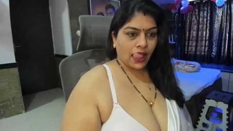 Media: A video of a South Asian woman with long black hair and medium skin tone, wearing a white tank top and gold necklace, sticking out her tongue, indoors near a grey chair and a table with balloons.