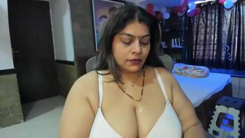 Media: Video of a South Asian woman with long black hair, fair skin, and large breasts, wearing a white bra, seated indoors in a room with a table, flowers, and balloons.