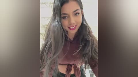 Media: Video of a young woman with long, wavy dark hair, wearing a pink button-up blouse tied at the waist, smiling directly at the camera, with a blurred, neutral-colored background.