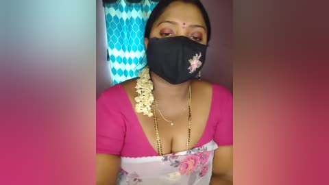 Media: Video of a South Asian woman in traditional attire, wearing a black mask, pink blouse, floral sari, and intricate jewelry. Background features a blue and white diamond-patterned curtain.