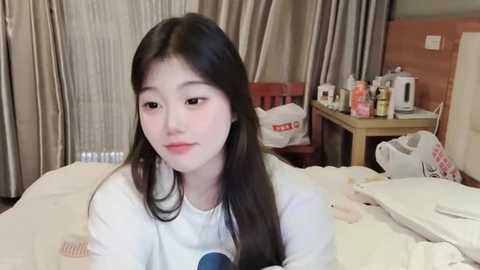 Media: Video of a young East Asian woman with long black hair, pale skin, and a neutral expression, wearing a white shirt. She sits in a messy bedroom with beige curtains, a table with toiletries, and a white bag.