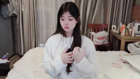 Media: Video of a young Asian woman with long black hair, wearing a white sweater, sitting on a bed, brushing her hair, in a bedroom with beige curtains, wooden furniture, and toiletries scattered around.