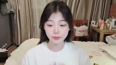 Media: Video of a young East Asian woman with fair skin and straight black hair, sitting in a dimly lit room. She wears a white shirt, and the background features a wooden table with toiletries and a pink chair.