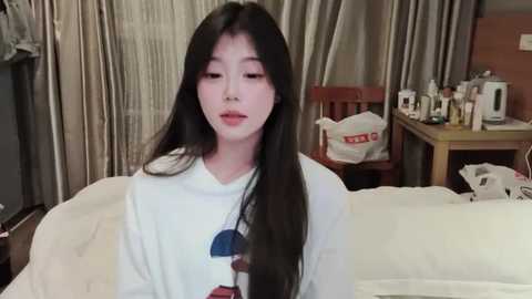 Media: A video of a young Asian woman with long black hair, fair skin, and soft makeup, wearing a white graphic tee, sitting on a bed in a cozy bedroom.