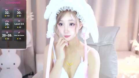 Media: A young Asian woman with fair skin and light blonde hair, wearing a frilly white hat and a pink bra, poses seductively in a softly lit bedroom.