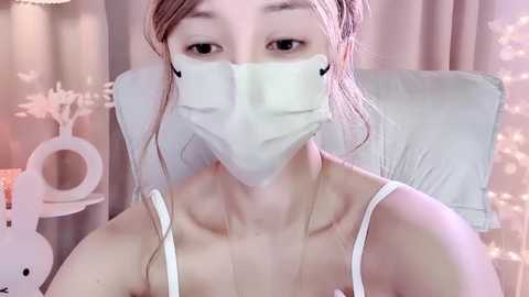 Media: Video of an Asian woman with light skin, wearing a white surgical mask and a white camisole, sitting on a white chair in a softly lit room with pastel-colored decor.