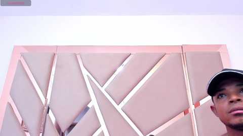 Media: Video of a young Black man with a shaved head wearing a black cap, looking upward at a modern, geometrically patterned wall with light pink and white triangles.