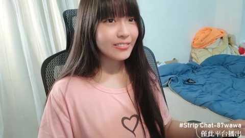 Media: Video of an East Asian woman with long black hair, wearing a pink T-shirt with a heart graphic, sitting on a black office chair in a messy bedroom with a blue blanket and clothes on the bed.