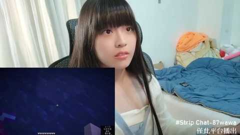 Media: Video of a young Asian woman with long black hair and fair skin, sitting on a chair in a messy bedroom, with a TV screen displaying a blue and white geometric pattern.