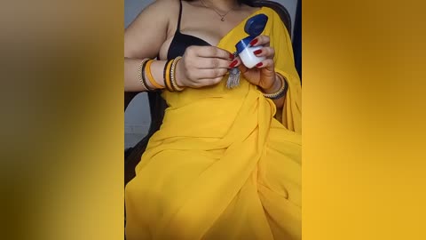 Media: Video of a woman in a yellow sari with black blouse, holding a blue and white spray can. She wears multiple bangles and a necklace.