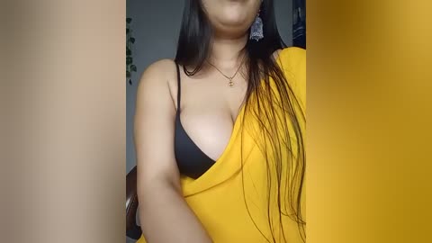 Media: Video of a woman with long black hair, wearing a yellow blouse with a black bra, and a gold necklace. The background is blurred.